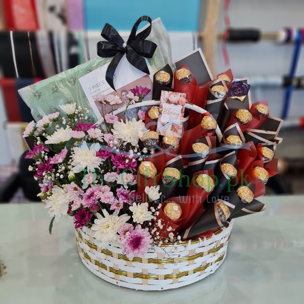 A gift basket of mixed color chrysanthemum flowers, Ferrero Rocher chocolates and Khadi ladies suits wrapped with a ribbon and a card.