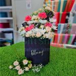 A bouquet of pink, white, and black flowers in a black box, on a green grassy surface with a blurred background of colorful fabrics.