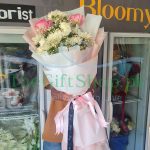 # A person holding a bouquet of pink roses and white chrysanthemums wrapped in white paper with a pink ribbon in front of a florist shop