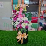 floral arrangement in a black box with a gold ribbon. The arrangement consists of pink roses, lilies, chrysanthemums, and baby’s breath. The vase is sitting on a green grass-like surface.