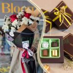 A gift set of a bouquet of white and red flowers and a box of assorted chocolates wrapped in brown and gold paper.