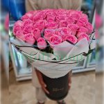 A large bouquet of bright pink roses in a black cylindrical box. The bouquet is wrapped in white paper.