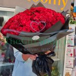 A person holding a large bouquet of 100 red roses wrapped in black paper in a flower shop.